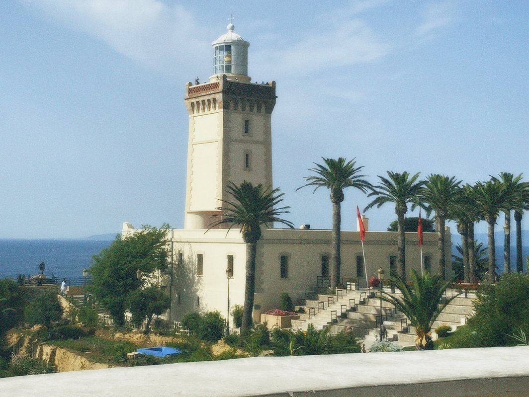 Photo of a lighthouse tower with a low building, palm trees and steps, with the sea behind.