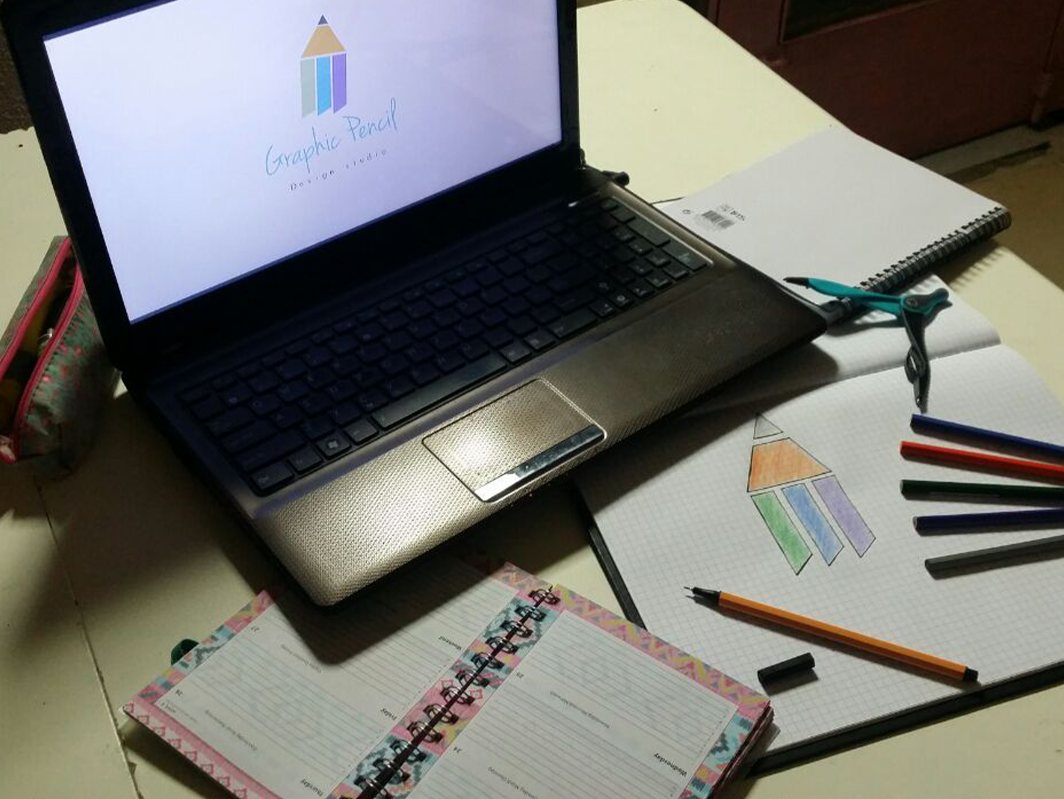 A laptop on a desk showing a coloured pencil logo, next to a squared notebook with the same drawing, coloured pencils and a pink planner.