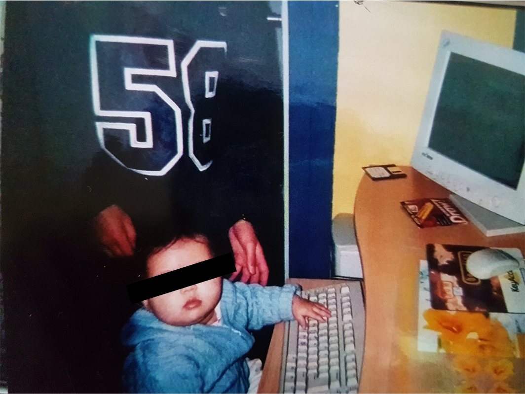 A baby at a desk next to a computer keyboard and monitor, with a black bar over the eyes.
