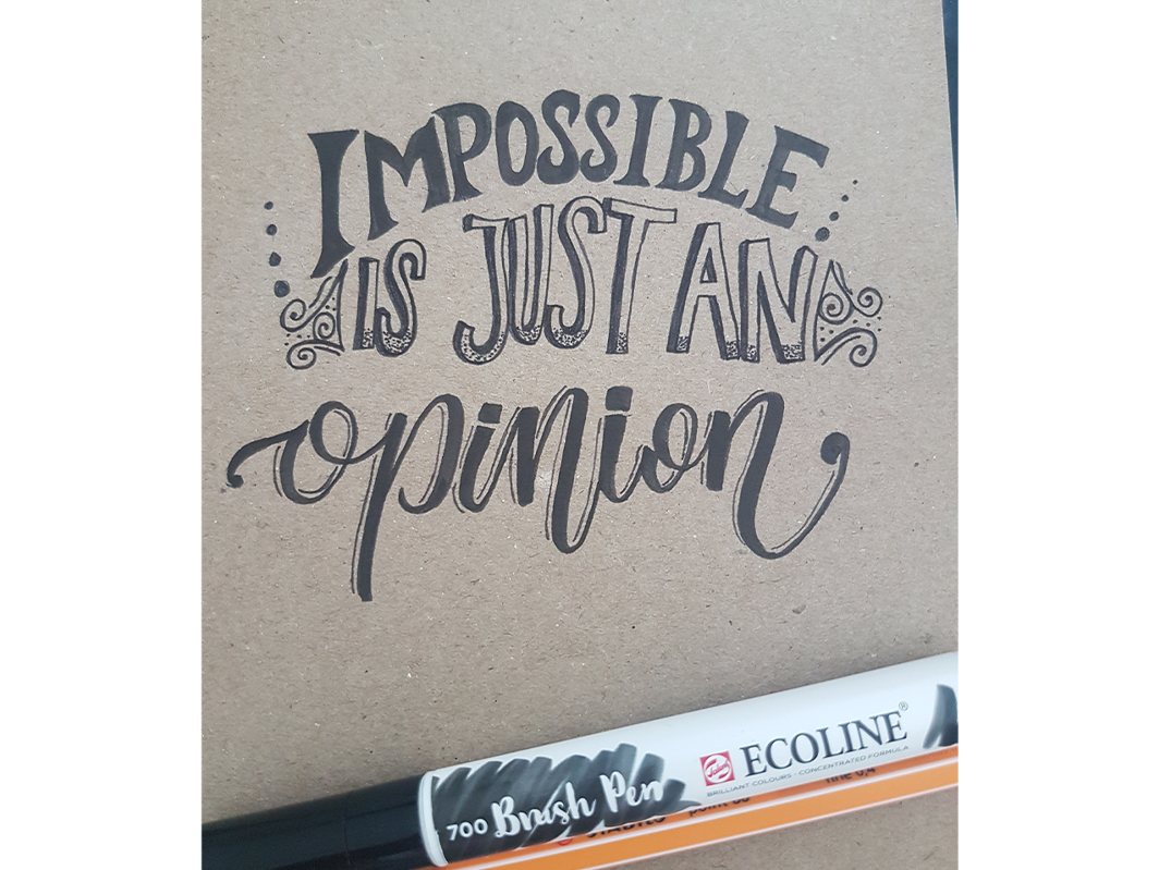 Hand lettering in black brush pen on brown paper: Impossible is just an opinion, with a brush pen lying across the corner.